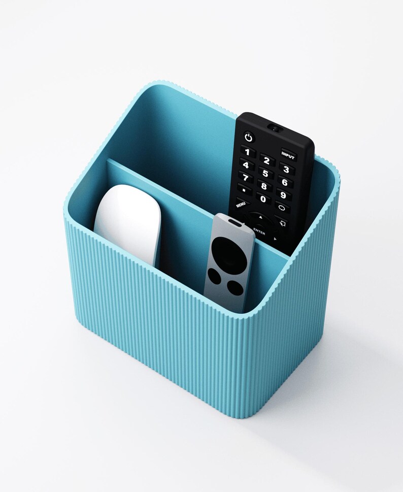 Puede incluir: Un organizador de escritorio azul claro con dos compartimentos. Un compartimento contiene un rat&oacute;n de ordenador blanco, mientras que el otro contiene un mando a distancia negro y un mando a distancia plateado. Dise&ntilde;ado para guardar material de oficina.