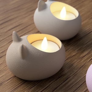 May include: Close-up of three ceramic candle holders shaped like rounded cat heads. The candle holders are light gray and lavender, with a textured surface. Each holds a glowing LED candle. The background is a wooden surface.