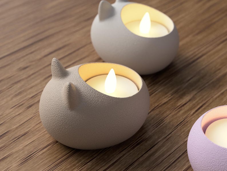 May include: Close-up of three ceramic candle holders shaped like rounded cat heads. The candle holders are light gray and lavender, with a textured surface. Each holds a glowing LED candle. The background is a wooden surface.