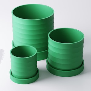 Op de afbeelding: Drie groene cilindrische bloempotten van verschillende maten. Elke pot heeft horizontale geribbelde details en staat op een bijpassende ronde basis. De potten hebben een levendige, effen groene kleur.
