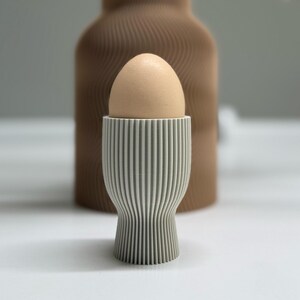 Pu&ograve; includere: Un uovo marrone chiaro &egrave; posto in un portauovo a righe grigie e bianche. Dietro il portauovo c'&egrave; un vaso marrone strutturato. Il portauovo ha un design a righe verticali. Lo sfondo &egrave; di colore neutro.