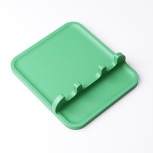 May include: A square, light green spoon rest with a raised edge and three curved slots for holding utensils. The rest is made of a solid, matte material and sits on a white background. The design is simple and modern.