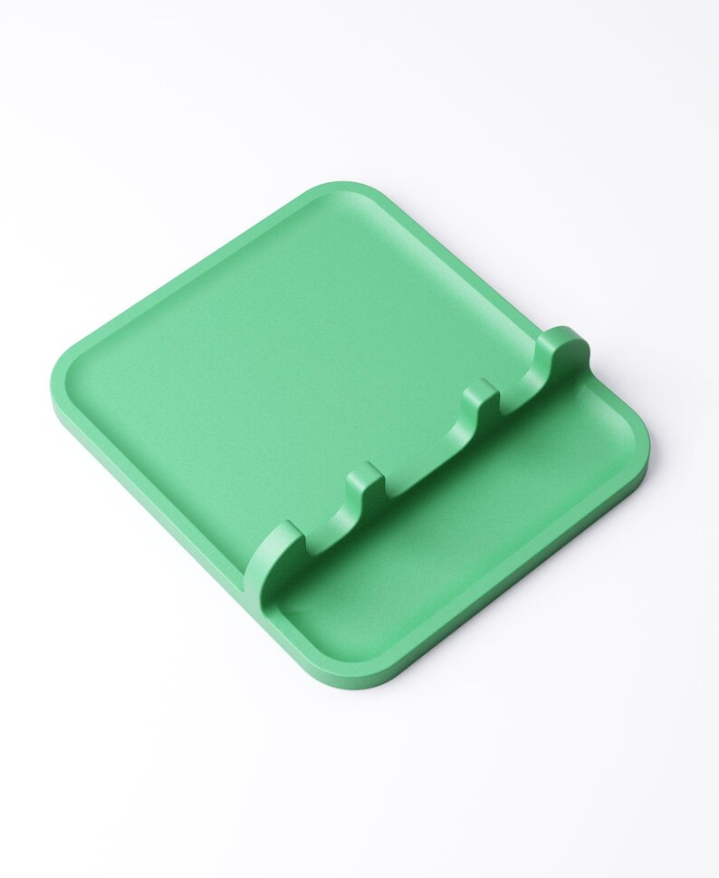 May include: A square, light green spoon rest with a raised edge and three curved slots for holding utensils. The rest is made of a solid, matte material and sits on a white background. The design is simple and modern.