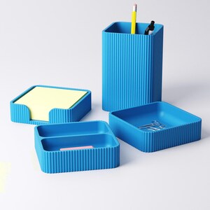 May include: A set of four matching blue desk organizers. The set includes a pencil holder with a yellow pencil and black marker, a square tray with paperclips, a small tray with a pink eraser, and a notepad holder with yellow sticky notes.