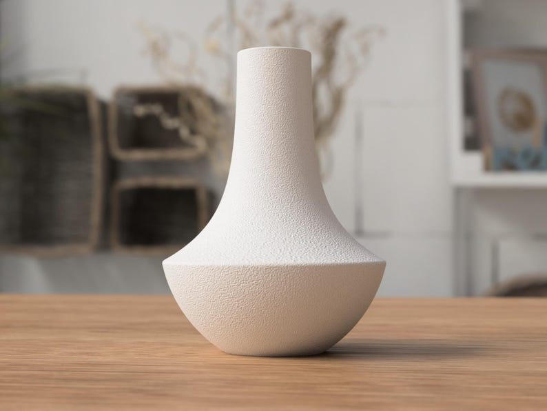 May include: A white, textured vase with a unique shape. The vase has a wide, rounded base that tapers to a narrow neck. The surface has a speckled texture. The vase is sitting on a wooden table.