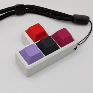 May include: A white rectangular holder with four colorful keycaps: red, black, magenta, and purple. The keycaps are arranged in a square. A black lanyard is attached to the holder, with a black clasp and a black cord.
