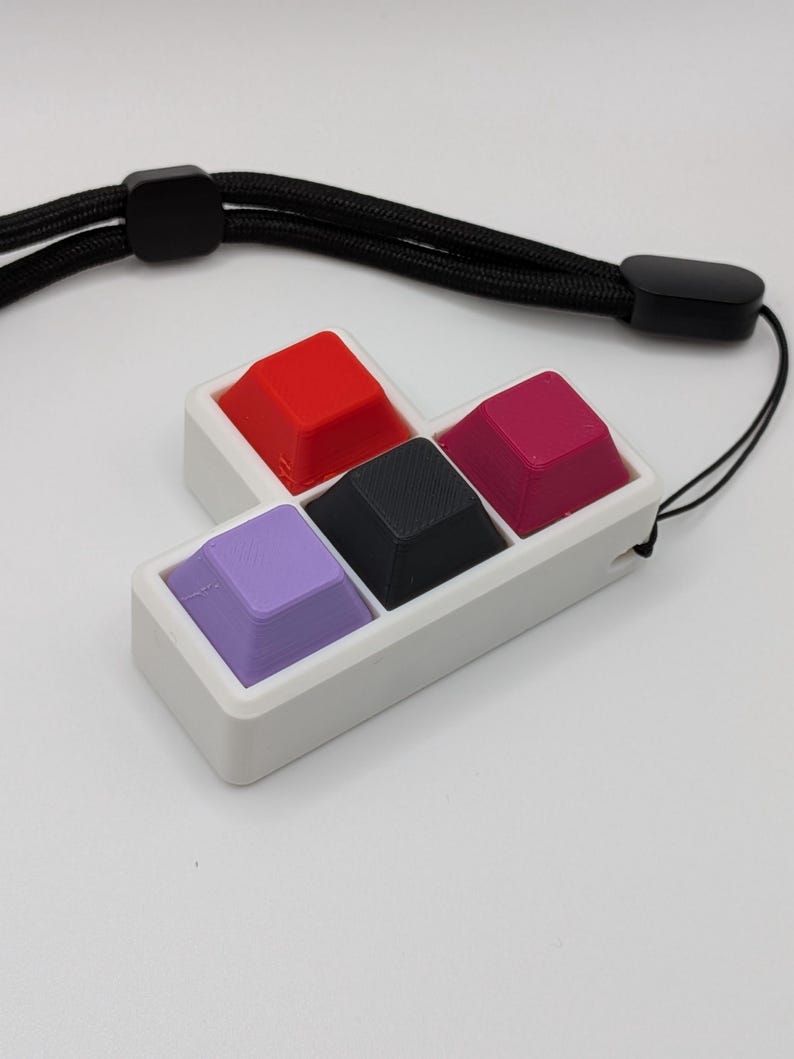 May include: A white rectangular holder with four colorful keycaps: red, black, magenta, and purple. The keycaps are arranged in a square. A black lanyard is attached to the holder, with a black clasp and a black cord.