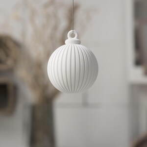 May include: A white, round Christmas ornament with vertical ribbed detailing. The ornament is suspended by a thin string, and has a loop at the top. The background is blurred, with hints of dried plants and a shelf.