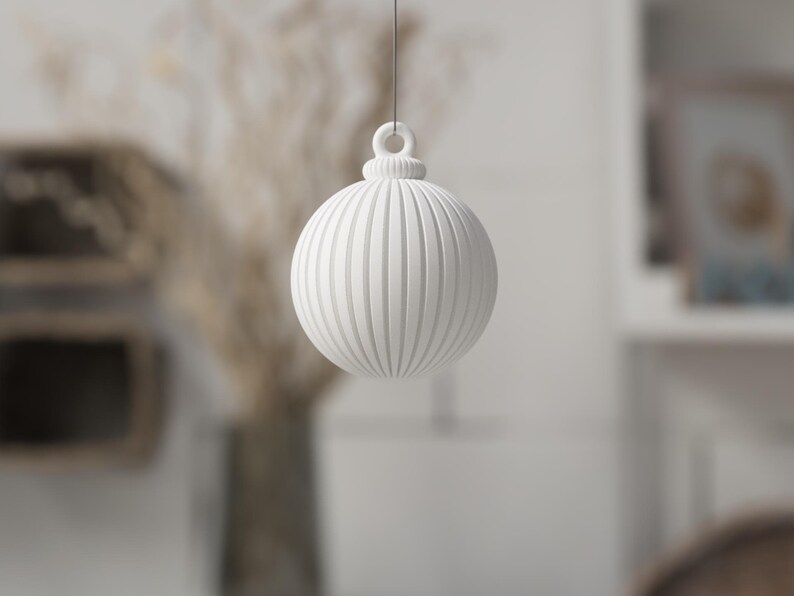 May include: A white, round Christmas ornament with vertical ribbed detailing. The ornament is suspended by a thin string, and has a loop at the top. The background is blurred, with hints of dried plants and a shelf.