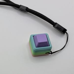 May include: A multi-colored, 3D-printed keycap with a lavender-colored square button. The keycap is set in a square frame with a gradient of colors, including blue, green, and pink. A black lanyard is attached to the keycap.