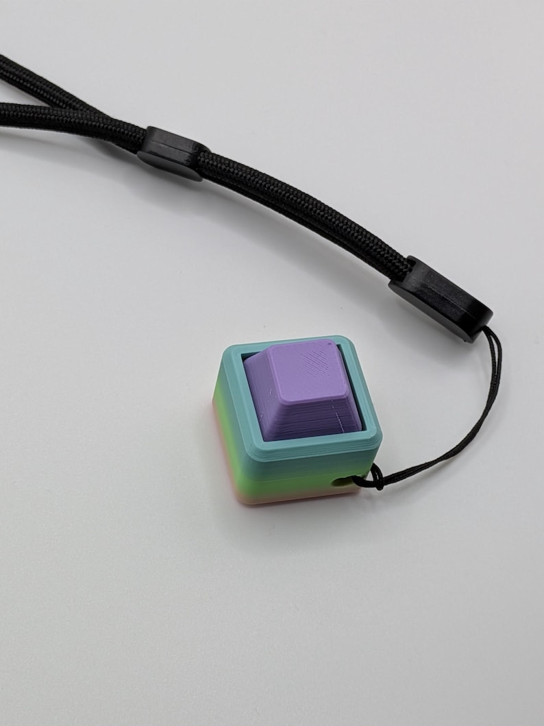May include: A multi-colored, 3D-printed keycap with a lavender-colored square button. The keycap is set in a square frame with a gradient of colors, including blue, green, and pink. A black lanyard is attached to the keycap.