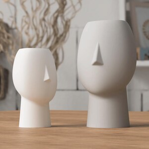 May include: Two white ceramic head-shaped vases with a minimalist design. The vases have a smooth, matte finish and a triangular nose detail. One vase is smaller than the other. They are placed on a wooden surface.