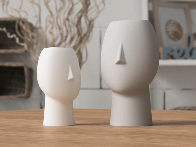 May include: Two white ceramic head-shaped vases with a minimalist design. The vases have a smooth, matte finish and a triangular nose detail. One vase is smaller than the other. They are placed on a wooden surface.