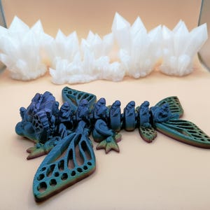 May include: A 3D-printed sculpture of a blue and green insect-like creature with intricate wing details. Above it are several white, crystalline formations. The objects are set against a neutral background, showcasing the detailed craftsmanship.