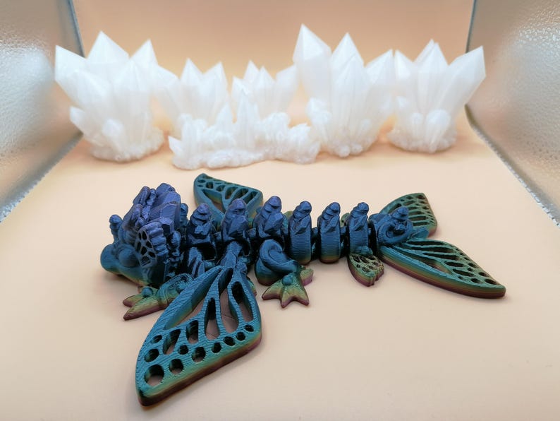 May include: A 3D-printed sculpture of a blue and green insect-like creature with intricate wing details. Above it are several white, crystalline formations. The objects are set against a neutral background, showcasing the detailed craftsmanship.