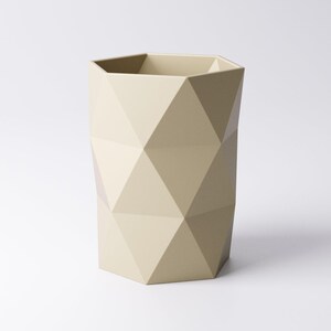 May include: A beige geometric wastebasket with a faceted design. The wastebasket has a hexagonal top and is made of a solid material. The surface is matte and the overall shape is a tall cylinder.