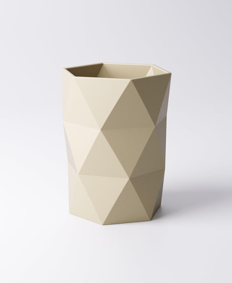 May include: A beige geometric wastebasket with a faceted design. The wastebasket has a hexagonal top and is made of a solid material. The surface is matte and the overall shape is a tall cylinder.