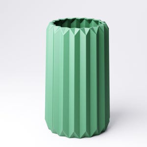 May include: A tall, green vase with a geometric, pleated design. The vase has a cylindrical shape with vertical ridges and a textured, angular rim. The color is a muted, pastel green, and the vase is set against a white background.