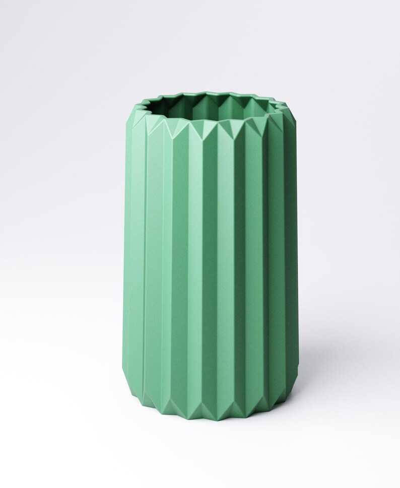 May include: A tall, green vase with a geometric, pleated design. The vase has a cylindrical shape with vertical ridges and a textured, angular rim. The color is a muted, pastel green, and the vase is set against a white background.