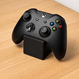 May include: Black Xbox controller resting on a black stand. The controller has colored buttons and two joysticks. The stand is a solid black square. The background is a light brown wooden surface.