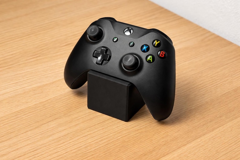 May include: Black Xbox controller resting on a black stand. The controller has colored buttons and two joysticks. The stand is a solid black square. The background is a light brown wooden surface.