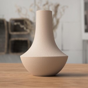 May include: A beige ceramic vase with a textured surface. The vase has a wide, rounded base that tapers to a narrow neck. The vase is sitting on a wooden table. The background is blurred, but appears to be a room with neutral tones.
