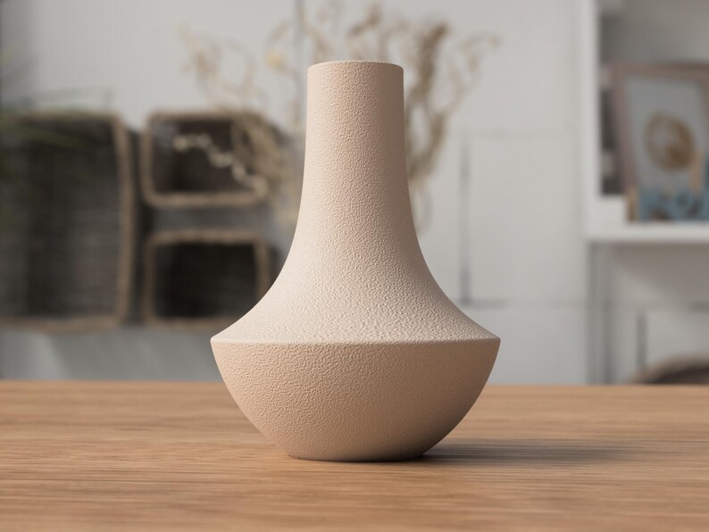 May include: A beige ceramic vase with a textured surface. The vase has a wide, rounded base that tapers to a narrow neck. The vase is sitting on a wooden table. The background is blurred, but appears to be a room with neutral tones.