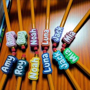 May include: A collection of yellow pencils with colorful name tags. The tags feature names like Amy, Roy, Noah, Luna, Jack, and Mia in various colors. Each pencil has a pink eraser.