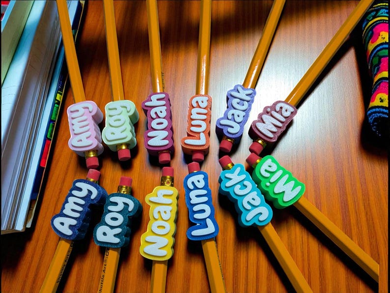 May include: A collection of yellow pencils with colorful name tags. The tags feature names like Amy, Roy, Noah, Luna, Jack, and Mia in various colors. Each pencil has a pink eraser.