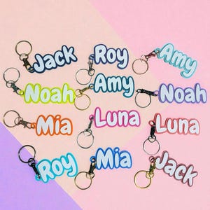 May include: A collection of personalized keychains with names like "Jack," "Roy," and "Amy" in various colors. Each keychain features a metal ring and clasp. The background is a two-tone pink and purple.