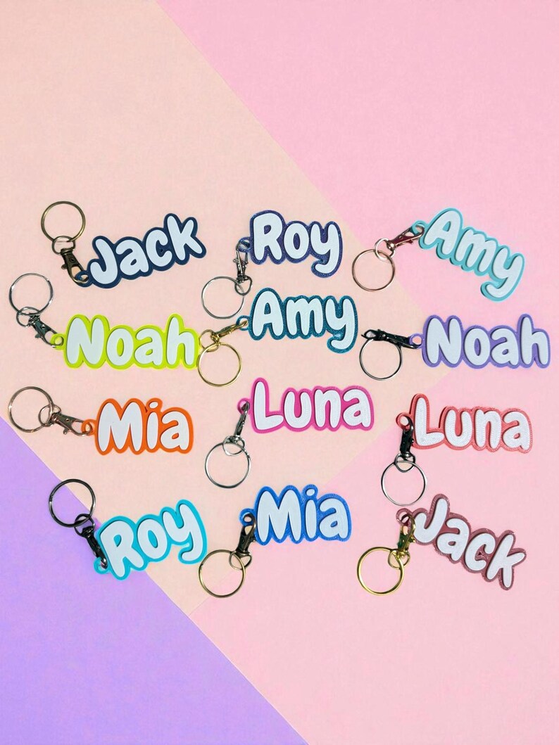 May include: A collection of personalized keychains with names like "Jack," "Roy," and "Amy" in various colors. Each keychain features a metal ring and clasp. The background is a two-tone pink and purple.