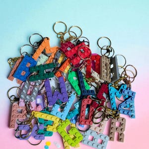 May include: A collection of colorful letter keychains made from interlocking plastic bricks. The keychains feature various colors, including red, blue, green, and orange, with gold and silver key rings. The background is a gradient of pink and blue.