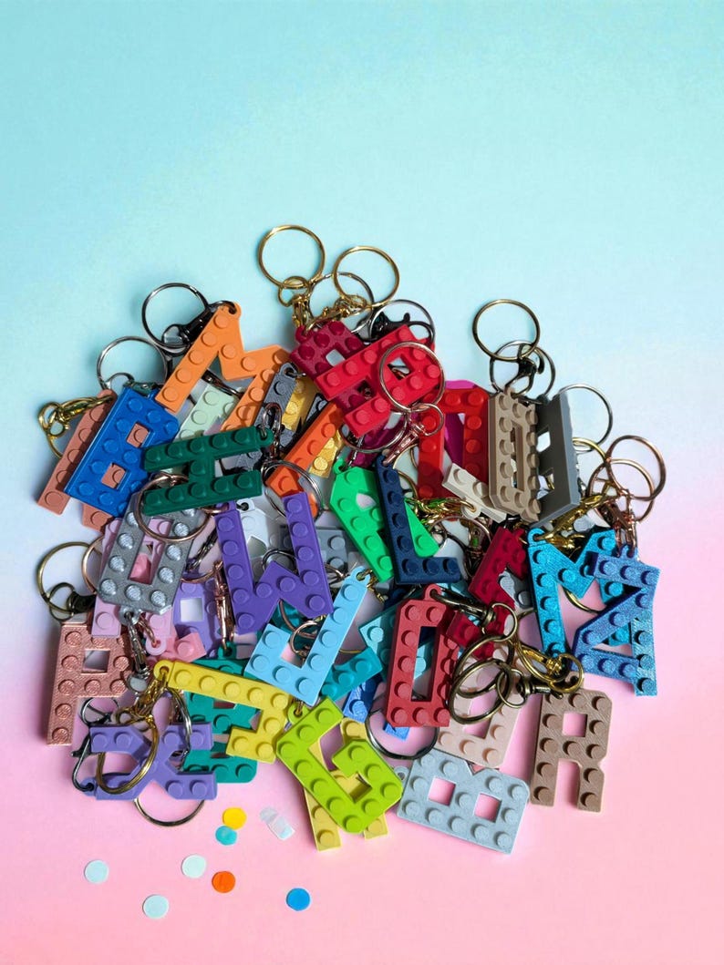 May include: A collection of colorful letter keychains made from interlocking plastic bricks. The keychains feature various colors, including red, blue, green, and orange, with gold and silver key rings. The background is a gradient of pink and blue.