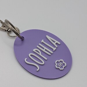 May include: A lavender oval keychain with the name "SOPHIA" in white, cursive lettering. A small white flower is also on the keychain. The keychain has a silver clasp and ring.