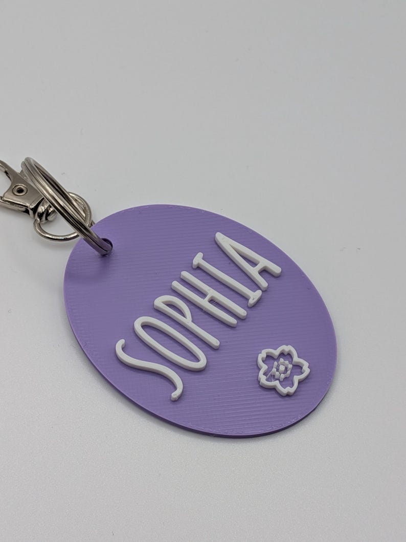 May include: A lavender oval keychain with the name "SOPHIA" in white, cursive lettering. A small white flower is also on the keychain. The keychain has a silver clasp and ring.