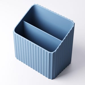 May include: A light blue desk organizer with a ribbed exterior and two interior compartments. The organizer has a rectangular shape with a slightly angled top edge. The color is a muted, pastel blue.