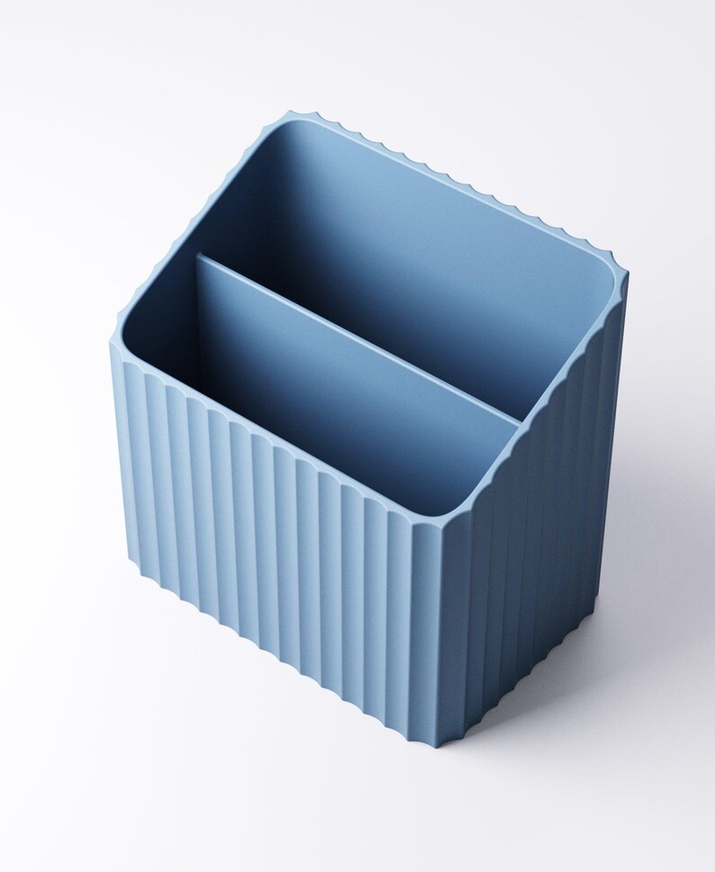 May include: A light blue desk organizer with a ribbed exterior and two interior compartments. The organizer has a rectangular shape with a slightly angled top edge. The color is a muted, pastel blue.