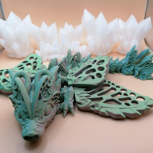 Pu&ograve; includere: Una scultura di drago dettagliata, stampata in 3D, in tonalit&agrave; di verde e blu, con intricate ali traforate e una testa dettagliata. Diverse strutture cristalline bianche sono sullo sfondo. &Egrave; un'opera d'arte fantasy.