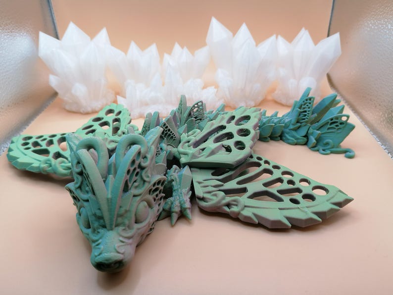 Pu&ograve; includere: Una scultura di drago dettagliata, stampata in 3D, in tonalit&agrave; di verde e blu, con intricate ali traforate e una testa dettagliata. Diverse strutture cristalline bianche sono sullo sfondo. &Egrave; un'opera d'arte fantasy.