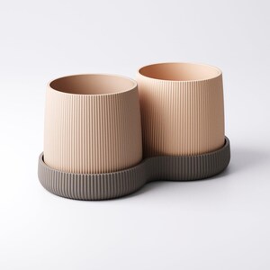 May include: Two beige cylindrical planters with vertical ribbed texture, sitting on a dark grey, wavy-edged base. The planters have a smooth interior and are suitable for indoor or outdoor use. The design is modern and minimalist.