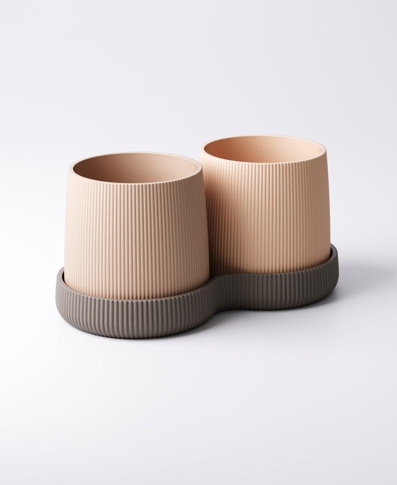 May include: Two beige cylindrical planters with vertical ribbed texture, sitting on a dark grey, wavy-edged base. The planters have a smooth interior and are suitable for indoor or outdoor use. The design is modern and minimalist.