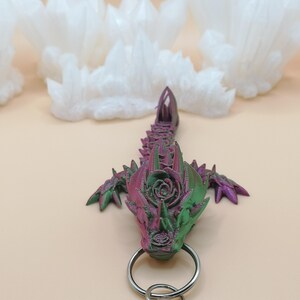 May include: A 3D-printed dragon keychain in shades of green and purple, with a silver keyring. The dragon has a detailed, textured design. The background features white, cloud-like shapes against a light beige surface.