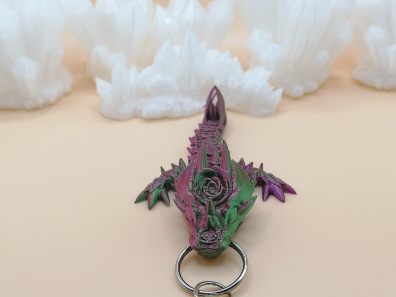 May include: A 3D-printed dragon keychain in shades of green and purple, with a silver keyring. The dragon has a detailed, textured design. The background features white, cloud-like shapes against a light beige surface.