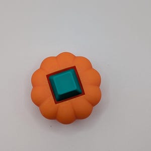 May include: An orange, flower-shaped object with a teal square in the center. The object has a red square border around the teal square. The object is likely a decorative item or a component of a larger design.