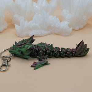 May include: A green and burgundy dragon keychain with a silver clasp and ring. The dragon is decorated with rose-like details. The background features white, crystalline formations.