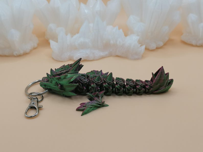 May include: A green and burgundy dragon keychain with a silver clasp and ring. The dragon is decorated with rose-like details. The background features white, crystalline formations.