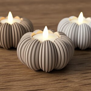 May include: Three decorative pumpkin-shaped candle holders with LED lights. The candle holders are in shades of gray and beige, with vertical ribbed detailing. The LED lights emit a warm, inviting glow, creating a cozy ambiance. The candle holders are placed on a wooden surface.