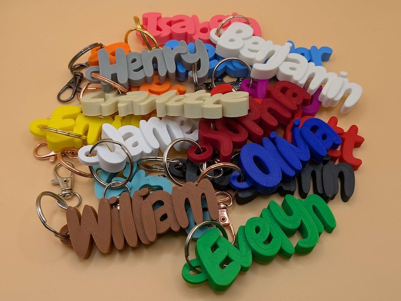 May include: A collection of colorful name keychains in various colors, including pink, blue, green, and brown. Each keychain features a different name in a 3D font, attached to a metal keyring. The keychains are scattered on a light orange surface.