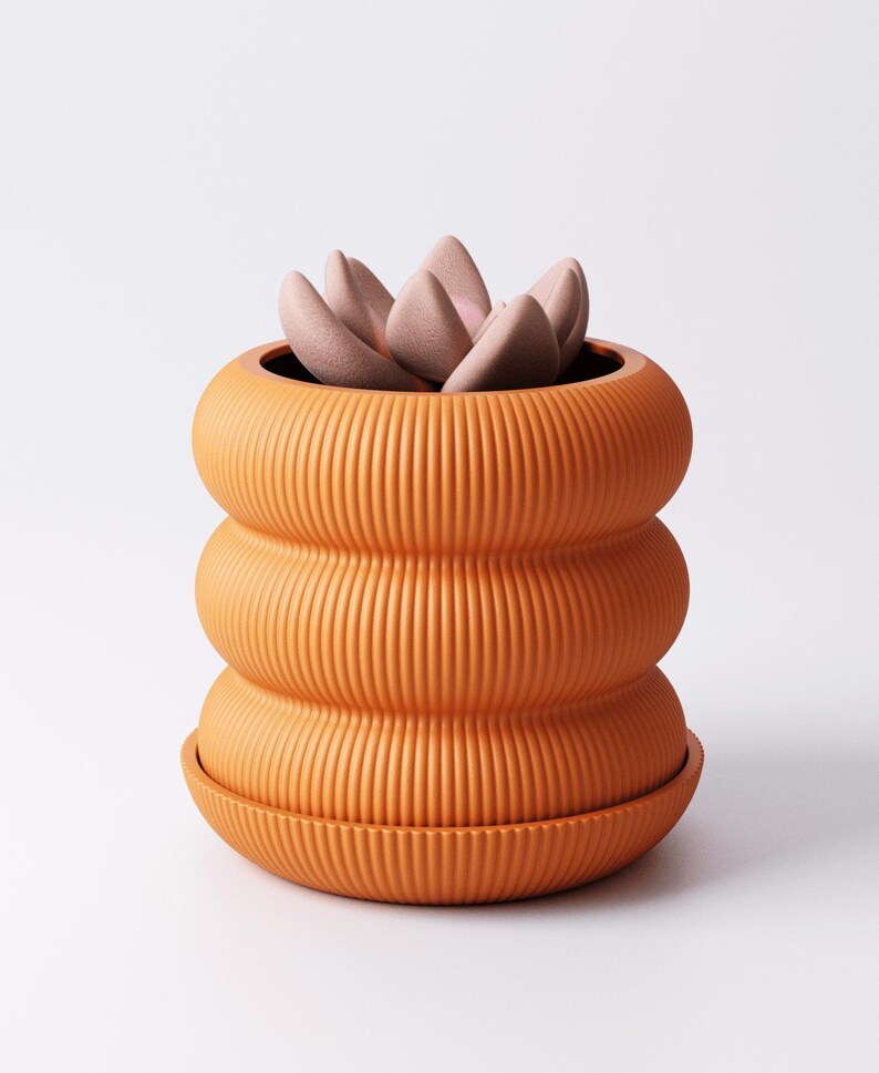 May include: A terracotta-coloured, ribbed ceramic plant pot with a matching saucer. The pot has a distinctive, stacked design and contains a succulent with pale pink leaves. The pot is a warm colour and would be a great addition to any home.