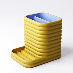 May include: A yellow desk organizer with a blue interior. The organizer has a rectangular shape with a series of horizontal ridges. A small tray is attached to the side. The design is modern and minimalist.